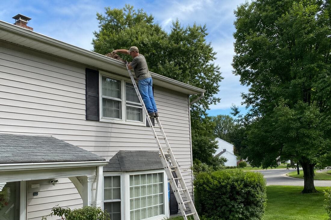 Homeowner climbing a tall extension ladder to clean gutters — a dangerous task that maintenance-free gutter guards eliminate